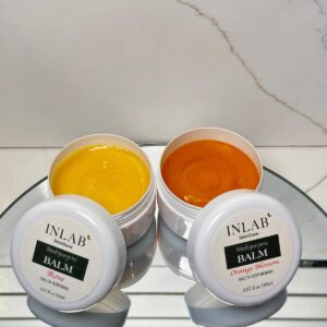 Two containers of Multi Purpose Balm one yellow and one orange, placed on a reflective surface against a minimalist background.