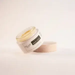 A clear glass jar of Deep Restore Night Cream with a golden lid, slightly tilted, beside a light wooden lid on a soft pastel background.