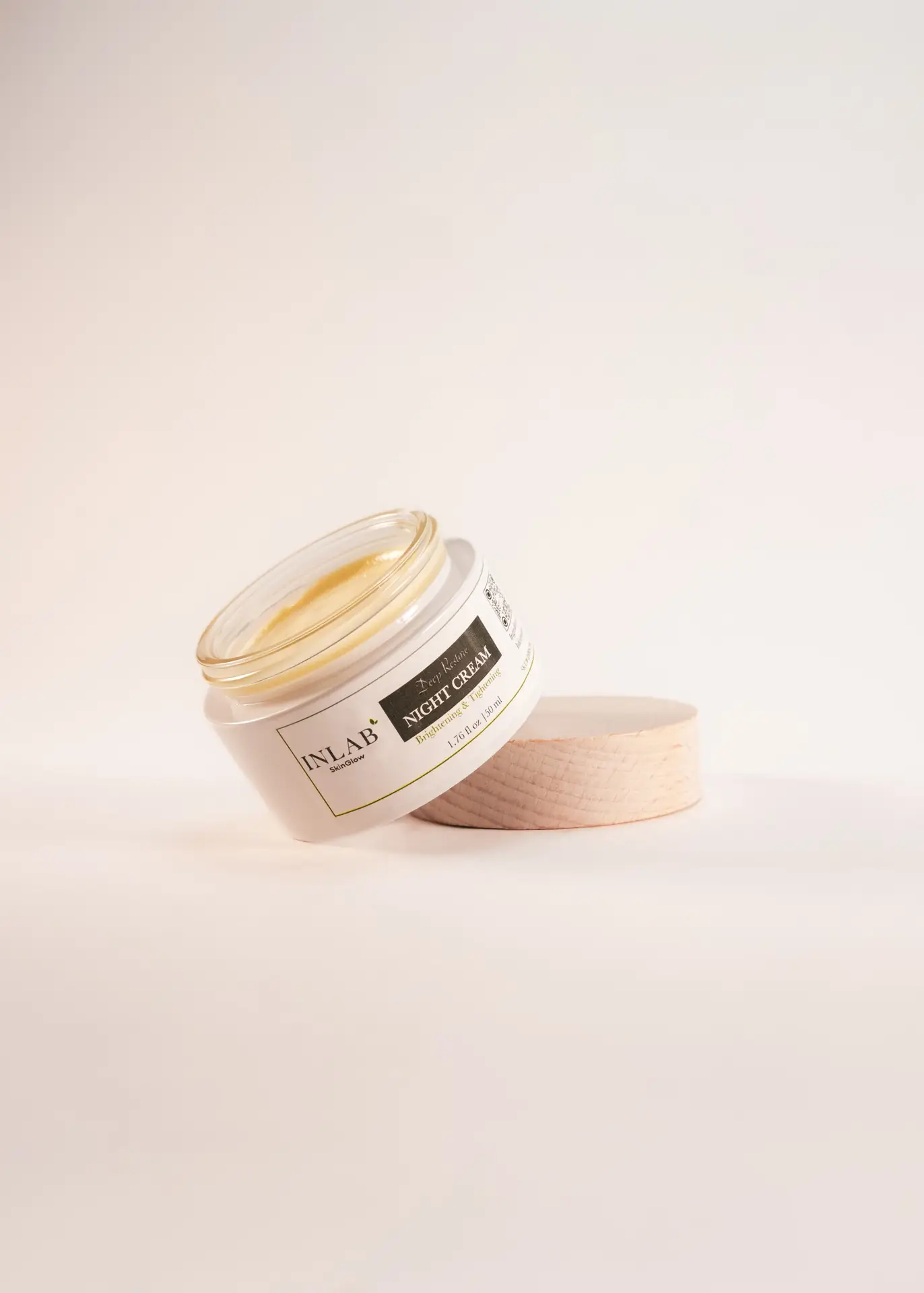 A clear glass jar of Deep Restore Night Cream with a golden lid, slightly tilted, beside a light wooden lid on a soft pastel background.